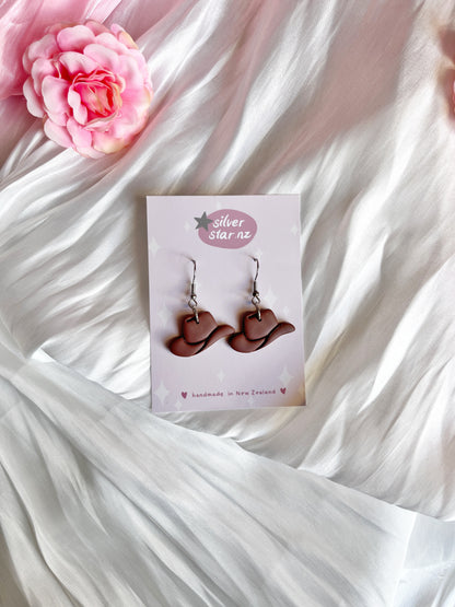 Cowboy Hat Polymer Clay Earrings by silverstar.nz feature a funky design on a card. Theyre crafted from handmade polymer clay with stainless steel hooks, set against white and pink satin fabric with a pink rose in the top left corner.