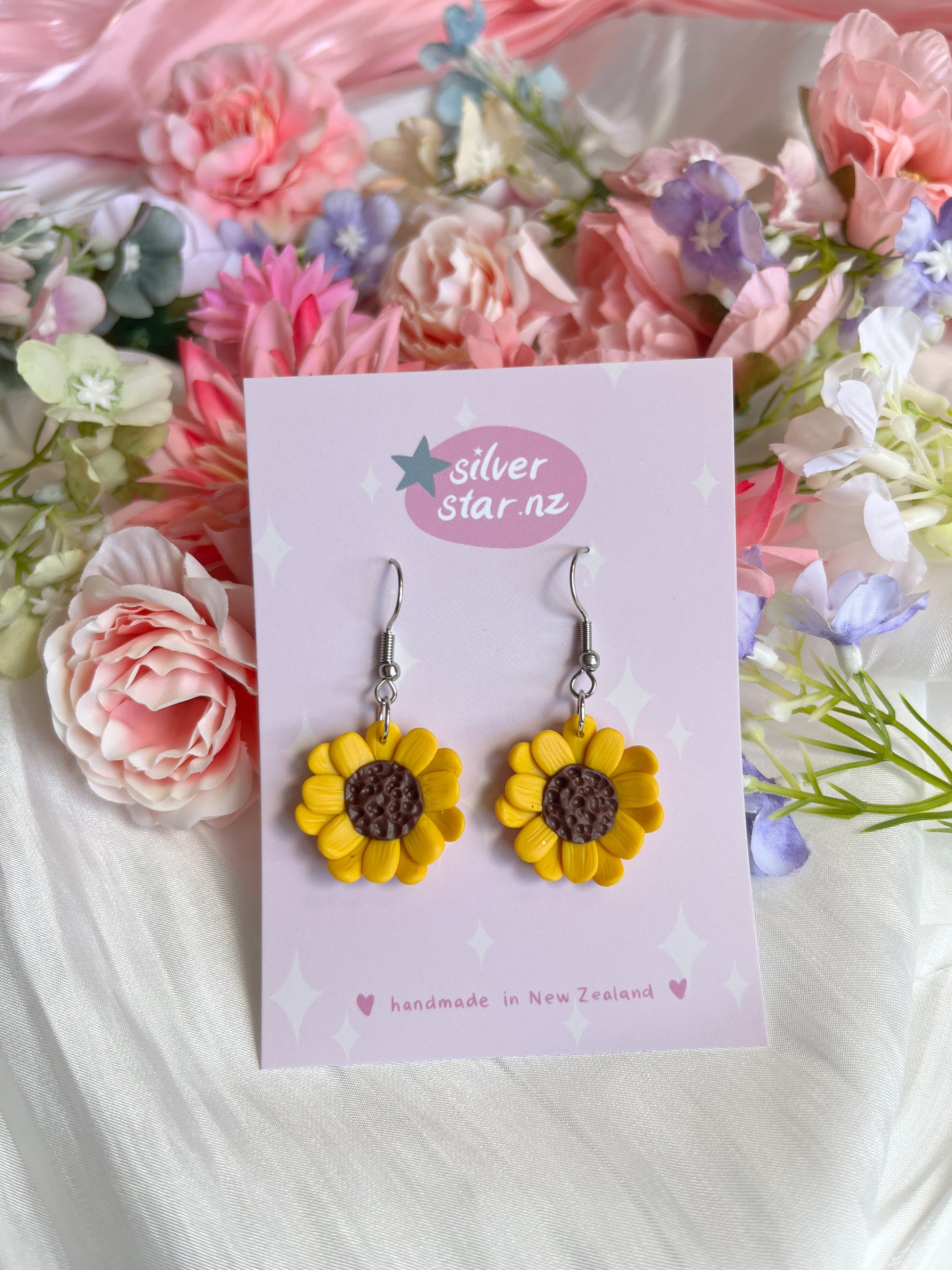 Handmade Sunflower Polymer Clay Earrings by silverstar.nz with brown centers and yellow petals are displayed on a card. The backdrop features pink, purple, and white flowers.