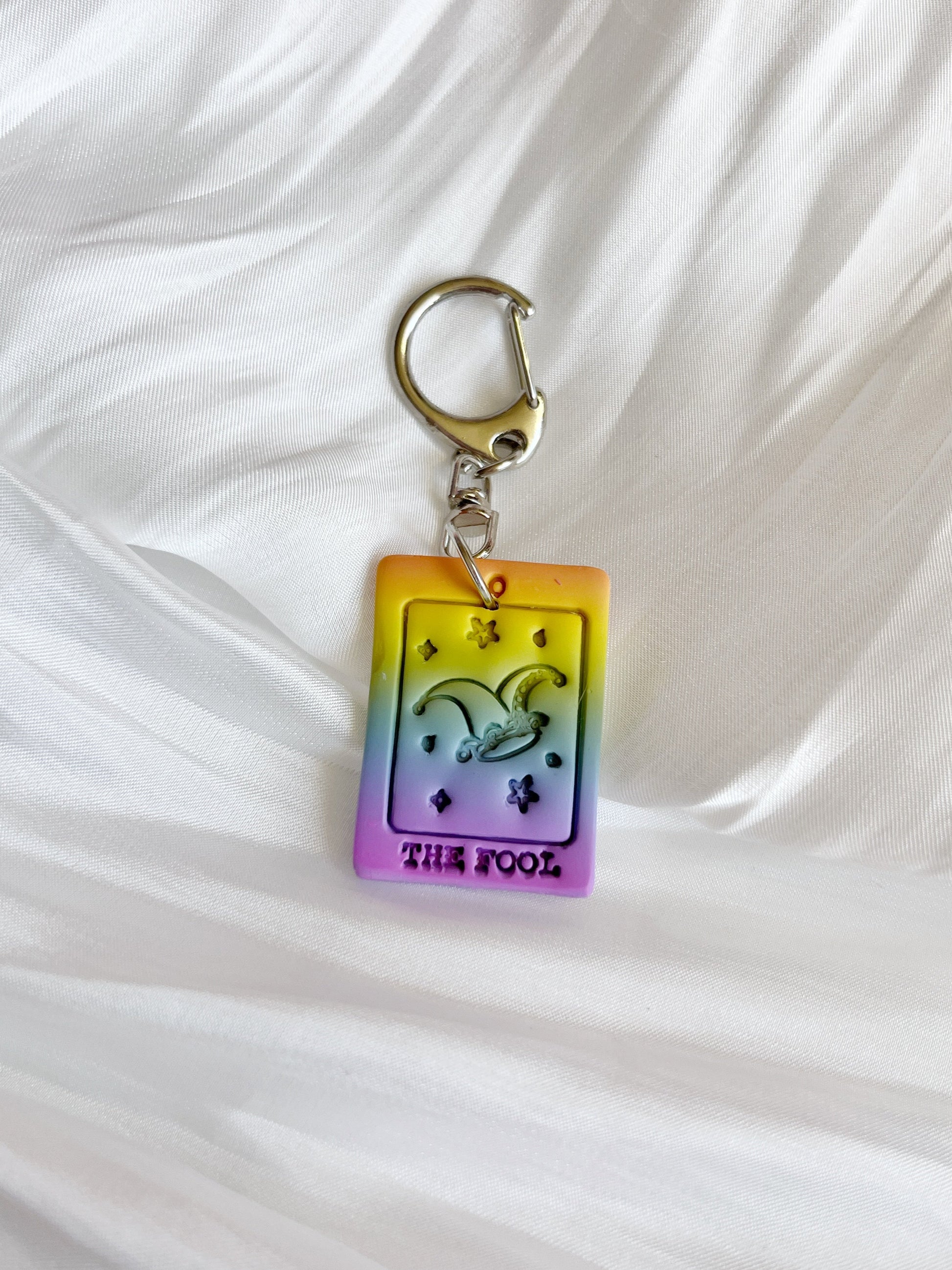 The Tarot Card Polymer Clay Keyring by silverstar.nz features a colorful The Fool tarot card design with a jester hat, showcasing vibrant yellow, orange, and purple hues against a white textured fabric background. Handcrafted in New Zealand, it has a unique funky look.
