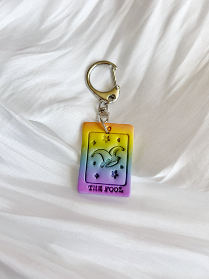 The Tarot Card Polymer Clay Keyring by silverstar.nz features a colorful The Fool tarot card design with a jester hat, showcasing vibrant yellow, orange, and purple hues against a white textured fabric background. Handcrafted in New Zealand, it has a unique funky look.