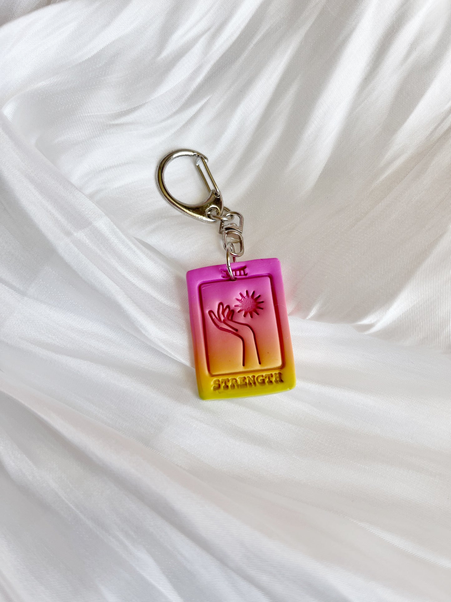 The Tarot Card Polymer Clay Keyring by silverstar.nz features a funky, colorful design with STRENGTH at the base, a hand holding a star, and rests on white fabric against a pink-yellow gradient. Its handcrafted in New Zealand using handmade polymer clay.