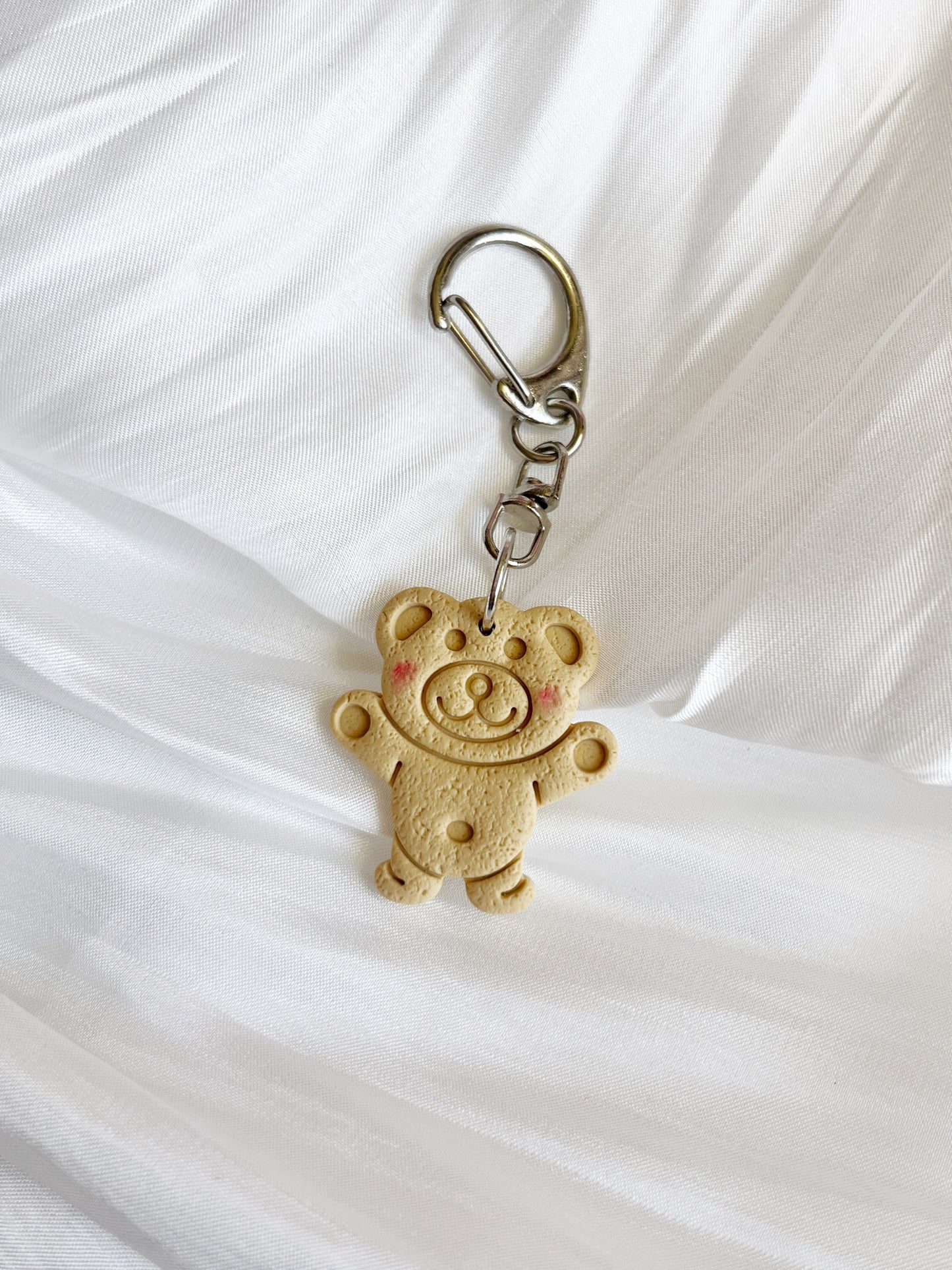 The Tiny Teddy Polymer Clay Keyring by silverstar.nz is a light brown teddy bear-shaped keychain with red cheeks and a smiling face, shown on a crumpled white fabric background.