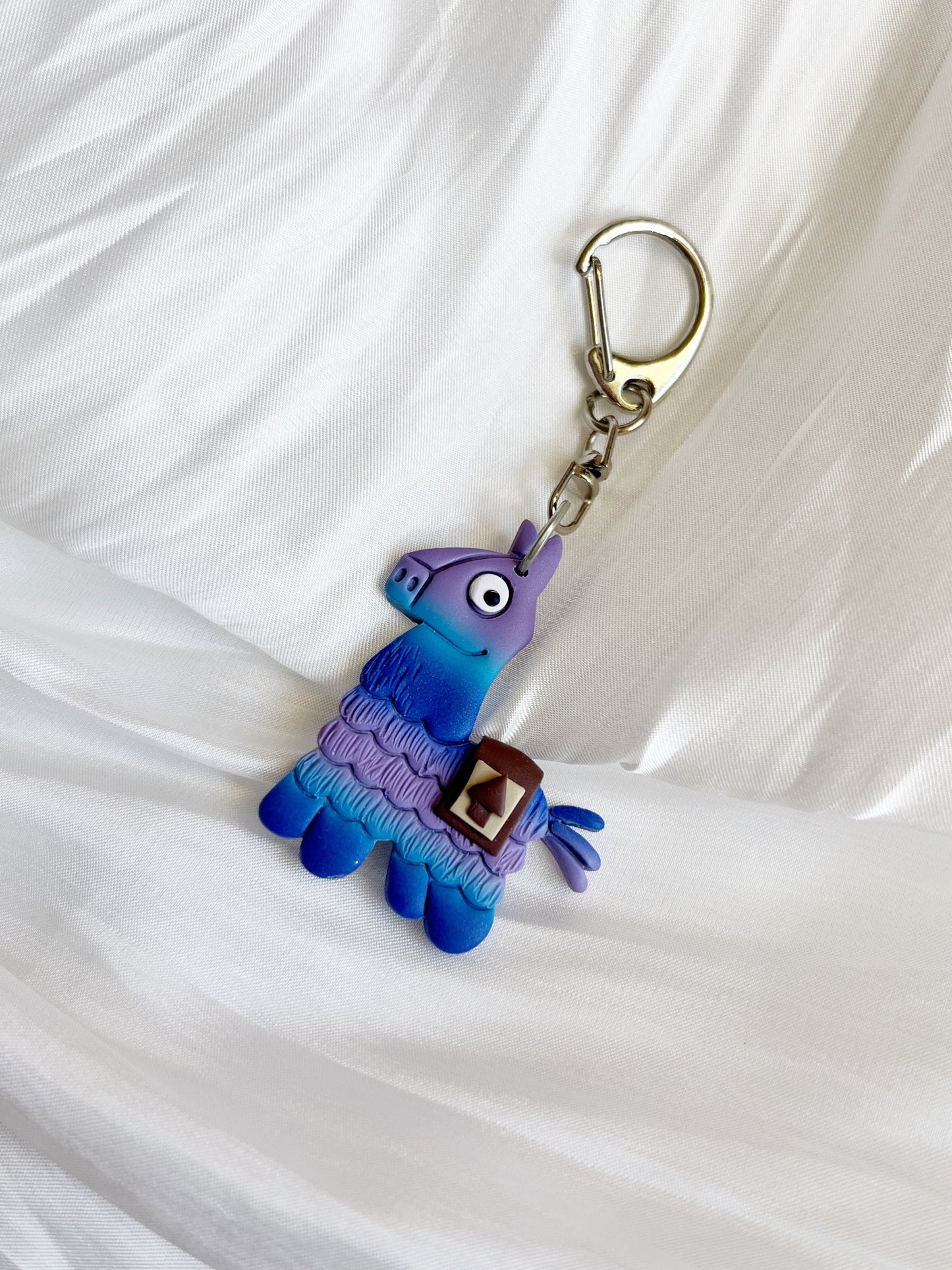 The Fortnite Llama Polymer Clay Keyring by silverstar.nz features a handmade, quirky llama in purple and blue tones on a textured white backdrop. This unique design is ideal for gifting.