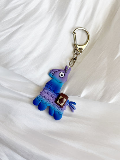 The Fortnite Llama Polymer Clay Keyring by silverstar.nz features a handmade, quirky llama in purple and blue tones on a textured white backdrop. This unique design is ideal for gifting.