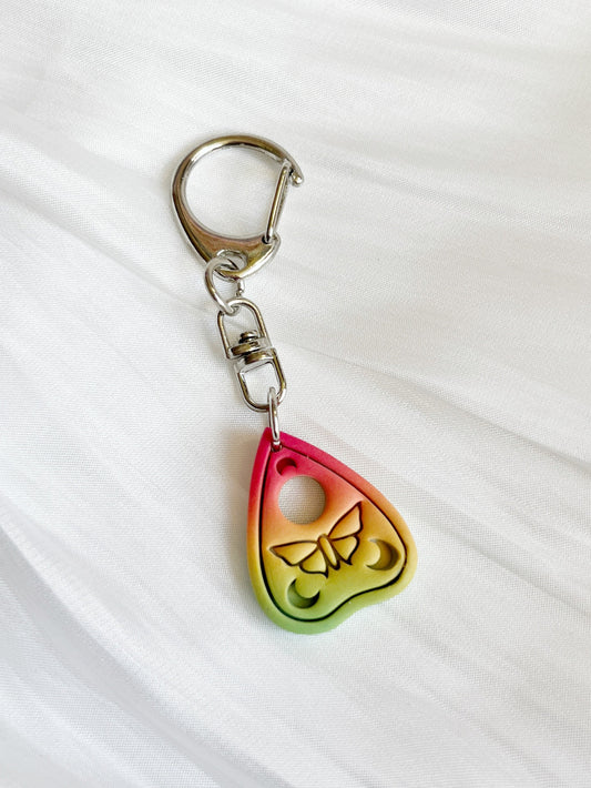 The Ouija Planchette Polymer Clay Keyring by silverstar.nz features a butterfly design with gradient colors, set against a white fabric background. This handcrafted, uniquely shaped keychain is made in New Zealand.
