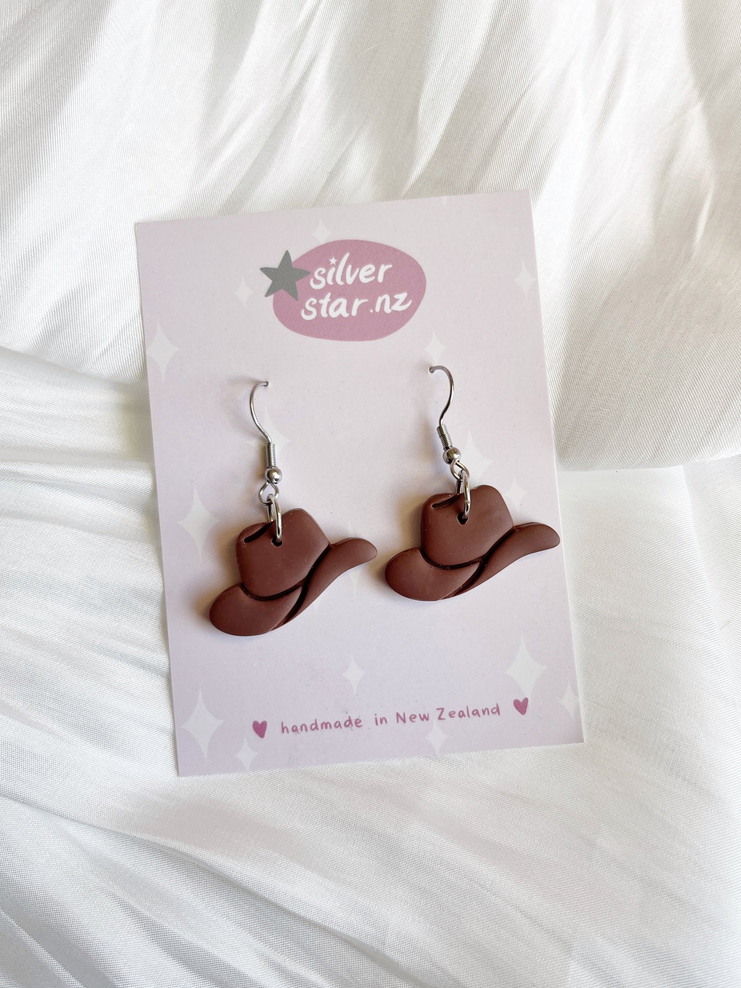 The Cowboy Hat Polymer Clay Earrings from silverstar.nz feature a funky brown cowboy hat design, handmade with stainless steel hooks. Crafted in New Zealand, they are elegantly displayed on a white fabric background.