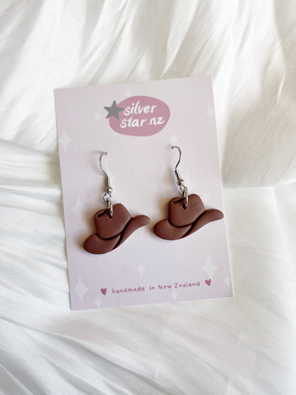The Cowboy Hat Polymer Clay Earrings from silverstar.nz feature a funky brown cowboy hat design, handmade with stainless steel hooks. Crafted in New Zealand, they are elegantly displayed on a white fabric background.