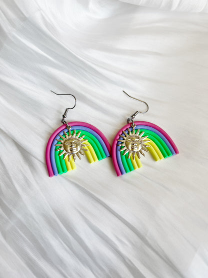 Silverstar.nzs Rainbow Arch Polymer Clay Earrings, featuring pink, blue, green, and yellow hues with silver sun charms, hang elegantly against a soft white fabric background for a unique design.