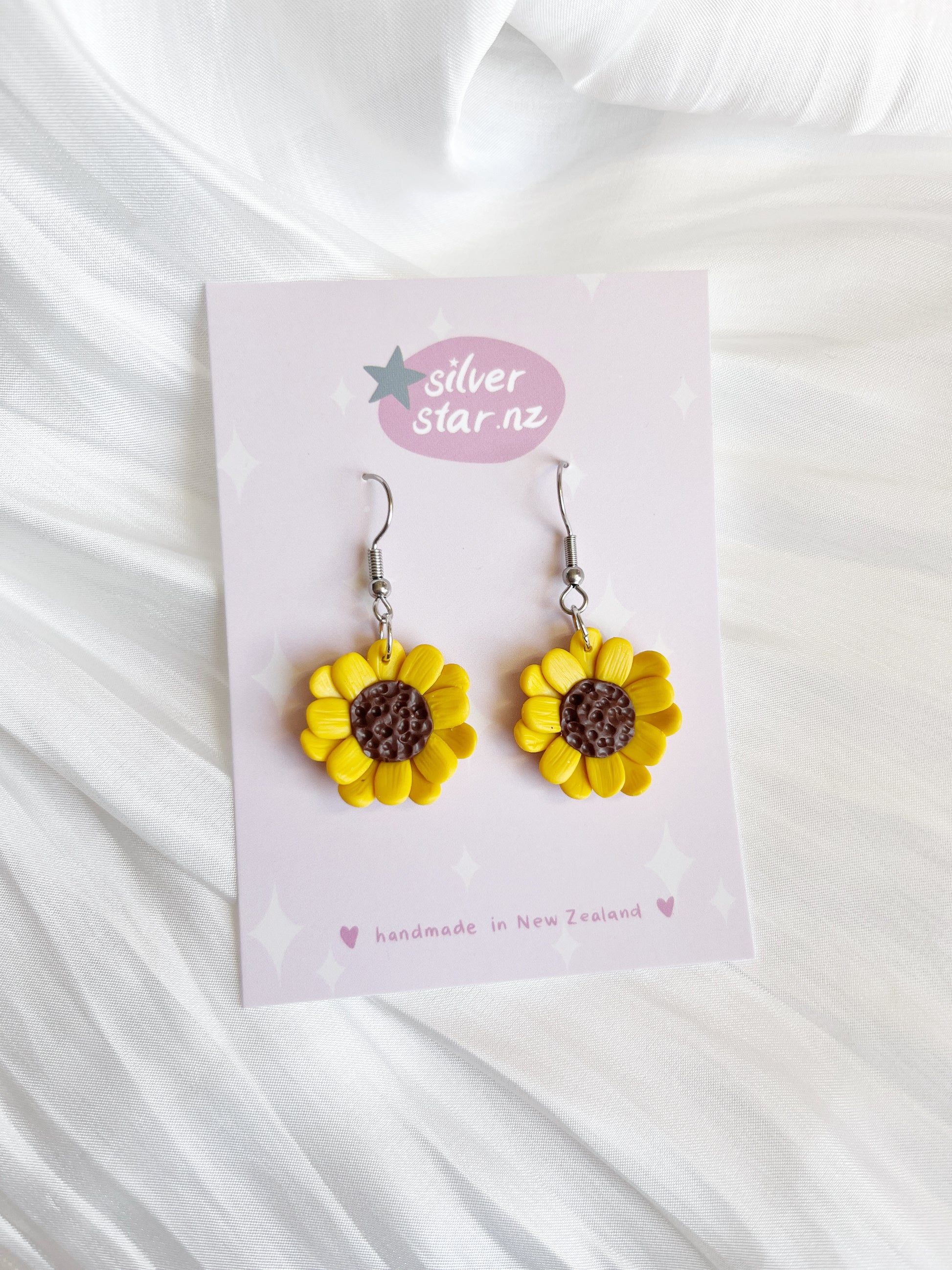 On a card labeled silverstar.nz, the Sunflower Polymer Clay Earrings, handmade in New Zealand with yellow petals and a brown center, are showcased against a soft white textured fabric.