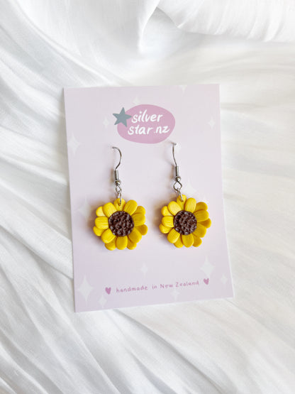 On a card labeled silverstar.nz, the Sunflower Polymer Clay Earrings, handmade in New Zealand with yellow petals and a brown center, are showcased against a soft white textured fabric.