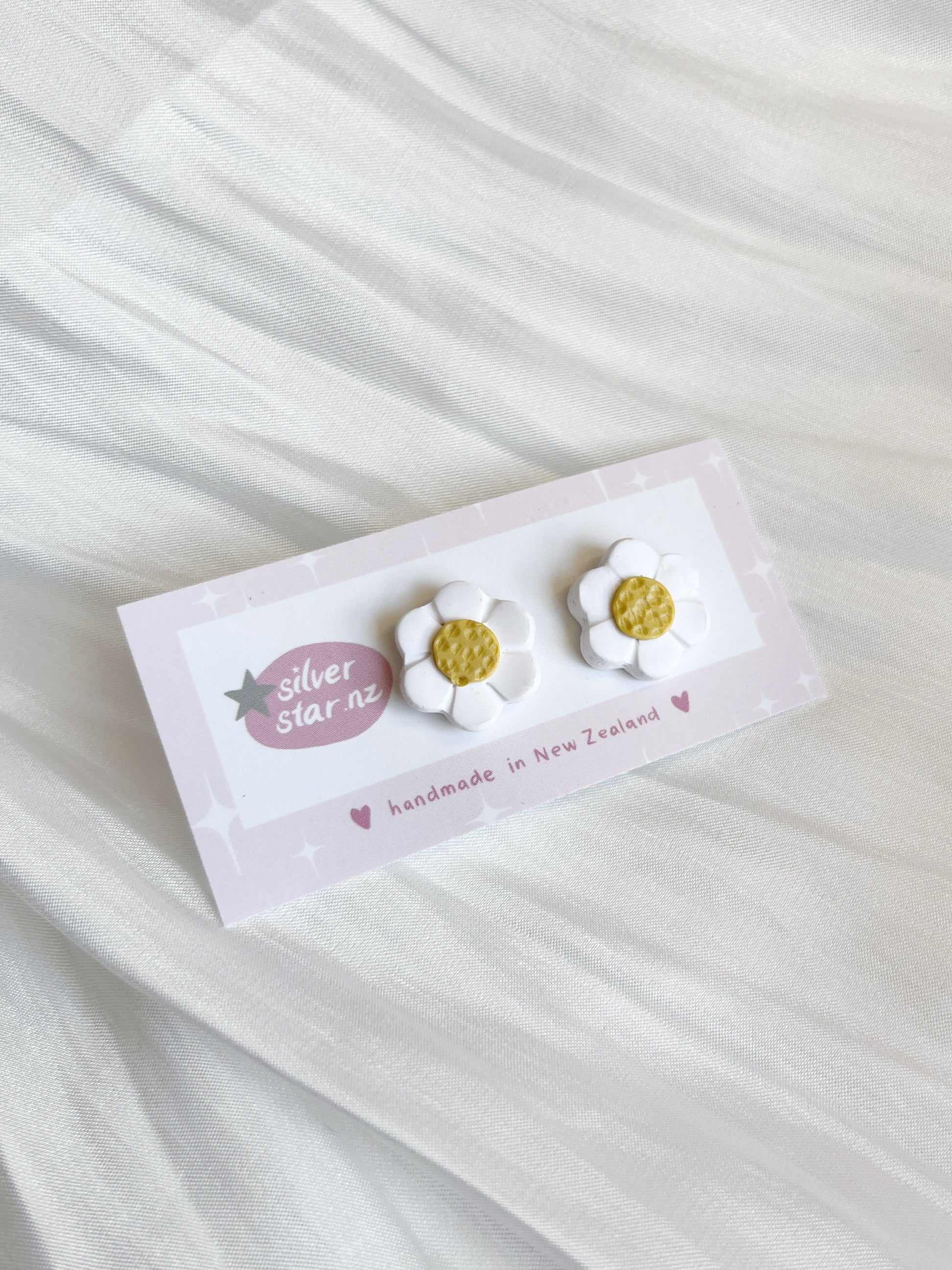 Displayed on a white textured fabric, silverstar.nz presents their White Daisy Polymer Clay Stud Earrings, featuring daisy-shaped earrings with white petals and yellow centers, proudly handmade in New Zealand.