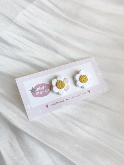 Displayed on a white textured fabric, silverstar.nz presents their White Daisy Polymer Clay Stud Earrings, featuring daisy-shaped earrings with white petals and yellow centers, proudly handmade in New Zealand.