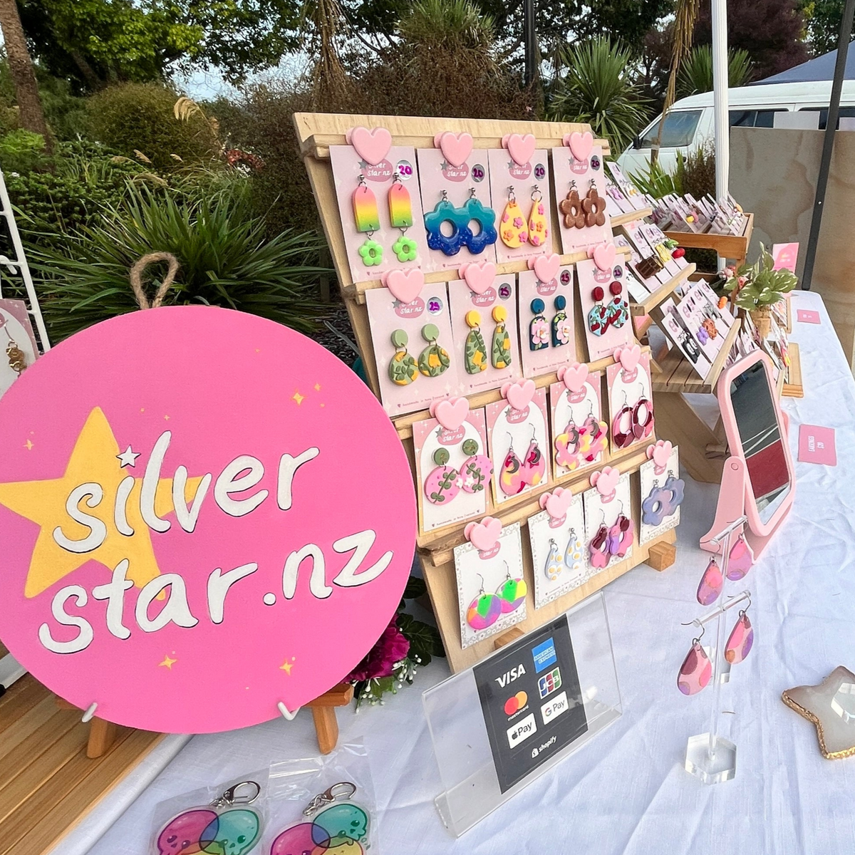 A display of colorful earrings on a wooden stand at an outdoor market stall. A round pink sign reads silver star.nz, and a small sign shows Visa and Mastercard logos. The table is covered with a white cloth.