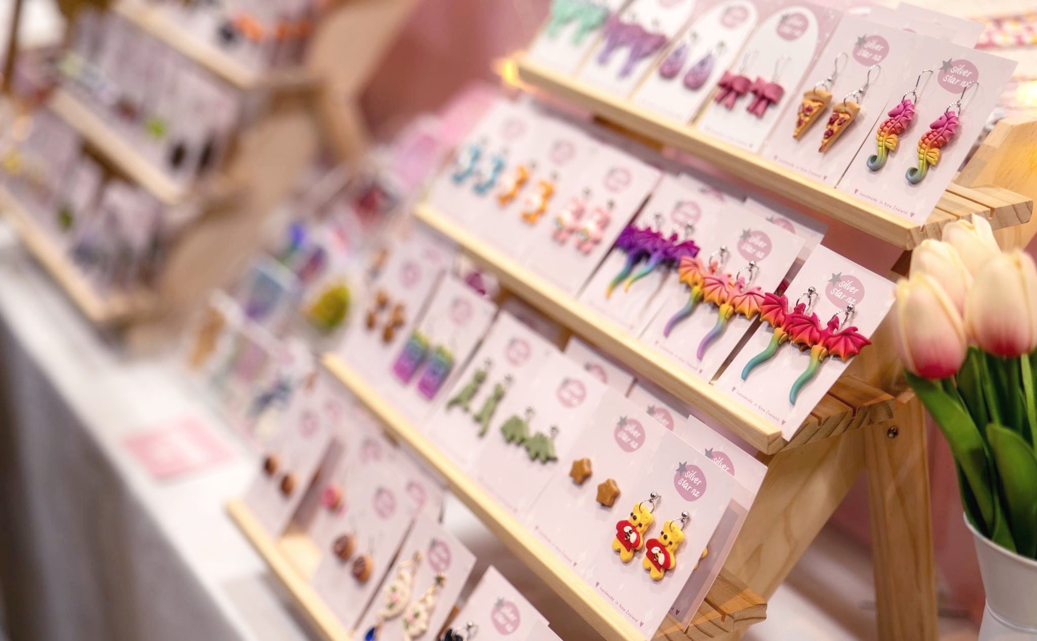 A display of colorful, handmade earrings on wooden stands. Each card has various designs, including flowers and food shapes. A bouquet of tulips is visible on the side.