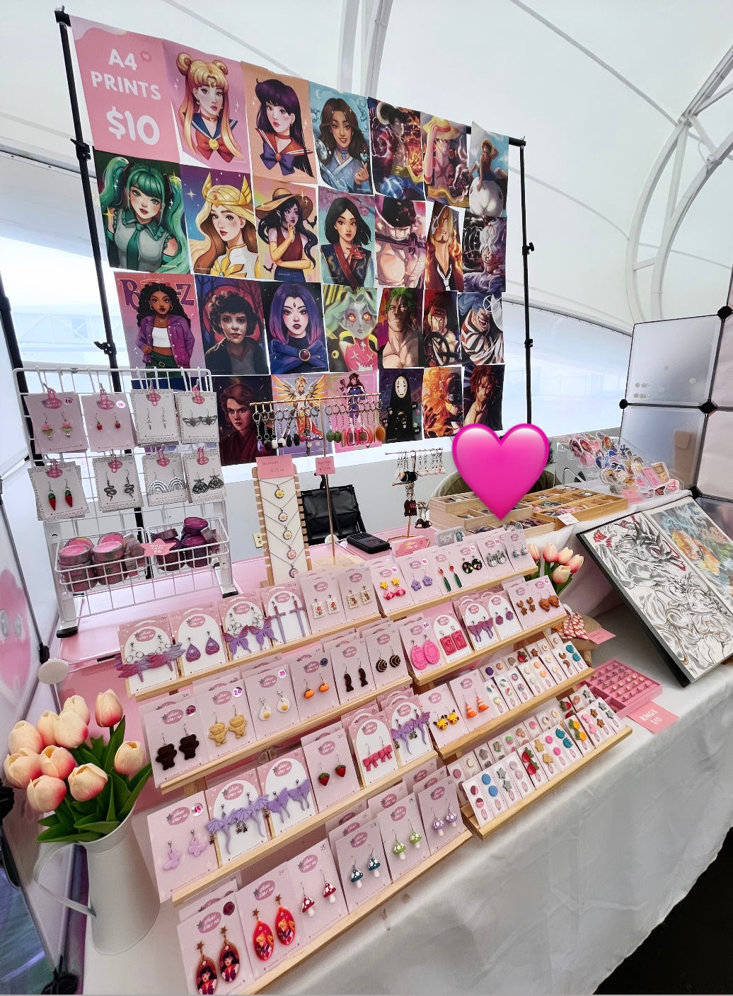 A booth displays various merchandise, including art prints, pins, keychains, and earrings. The setup features a colorful backdrop with illustrations of diverse characters. The table is neatly organized, highlighting the whimsical and vibrant items.