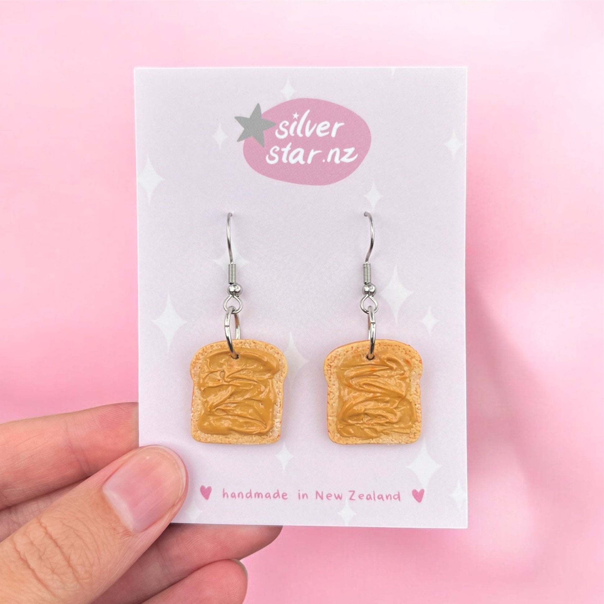 A hand displays a card featuring the "Peanut Butter on Toast Polymer Clay Earrings," designed with silver hooks to mimic peanut butter toast. The card, set against a pink background, bears the brand name "silverstar.nz" and declares "handmade in New Zealand.