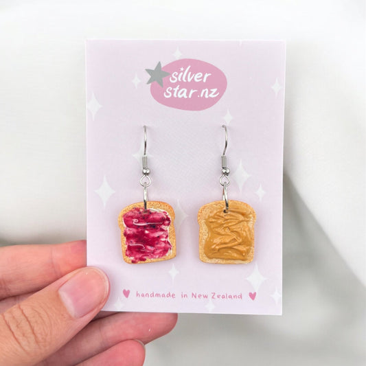 A hand holds a card displaying the Peanut Butter and Jelly Toast Polymer Clay Earrings. Each earring resembles a slice of bread, with one featuring red jelly and the other peanut butter. The earrings are equipped with stainless steel hooks, while the card bears the brand name silverstar.nz and notes that they are handmade in New Zealand.