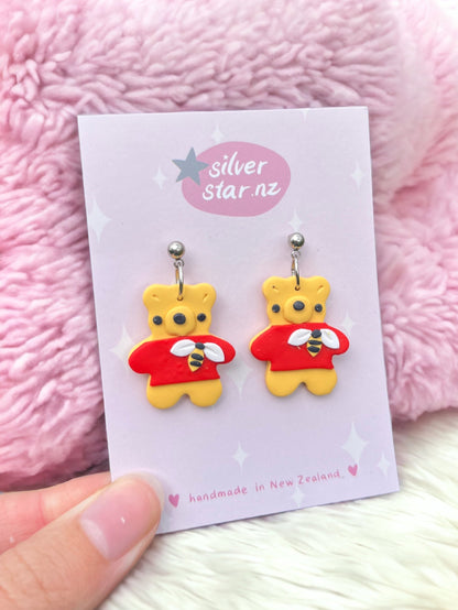 The Honey Bear Polymer Clay Earrings, uniquely designed as bears in red sweaters, are showcased on a card labeled silverstar.nz against a pink fluffy backdrop, with a hand delicately holding them at the bottom.