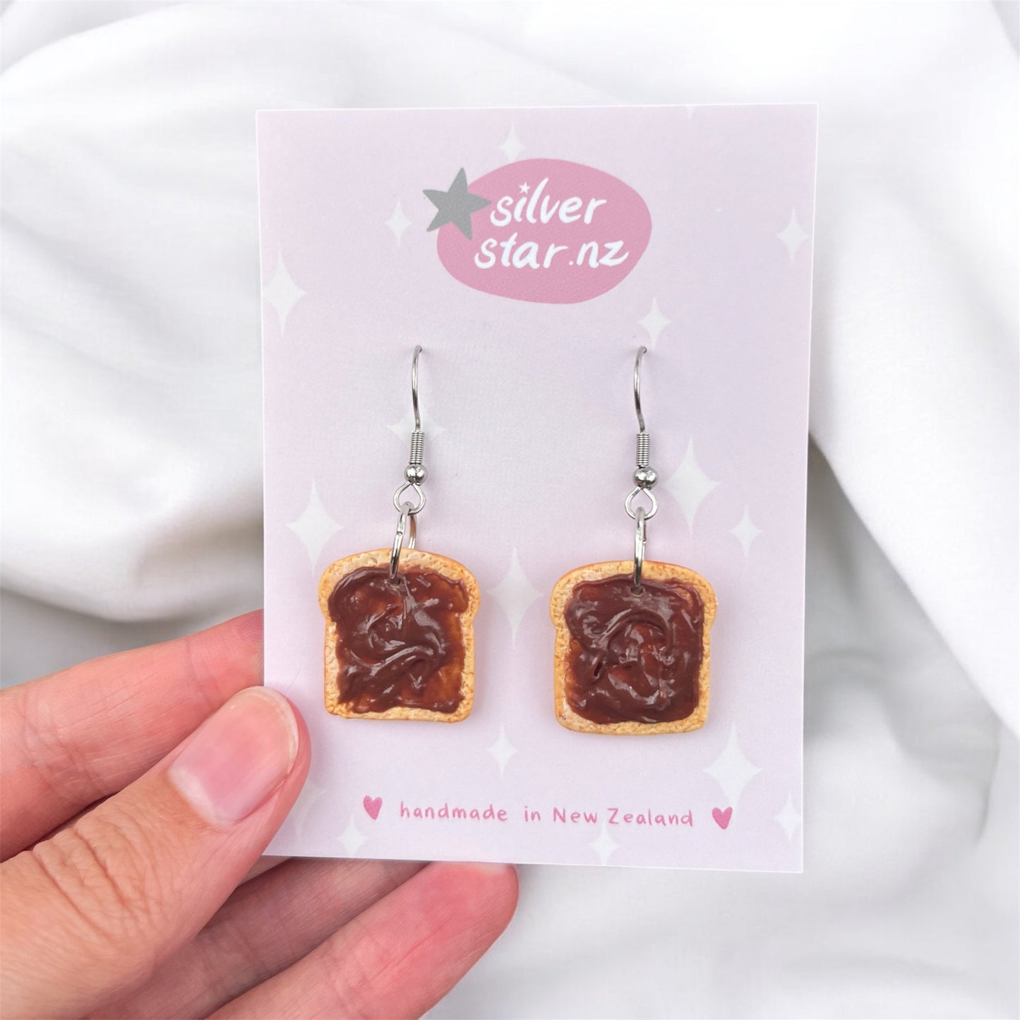 A hand holds a card showcasing distinct funky earrings shaped like slices of toast with a brown spread. The card reads "silverstar.nz" and "handmade in New Zealand." These Nutella Toast Polymer Clay Earrings are handmade and feature elegant silver hooks, adding a playful touch.