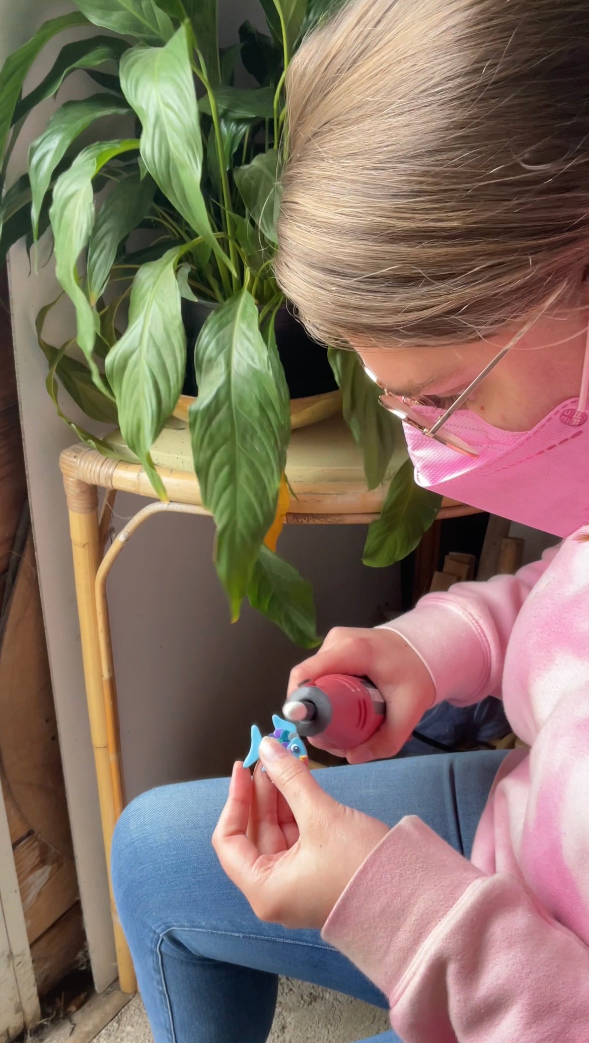 Sam holding a dremel in her hand and a handmade rainbow fish polymer clay earring in the other. Sam is sanding the edges of the fish earring to make the edges smooth and tidy. There is a green plant in the background.