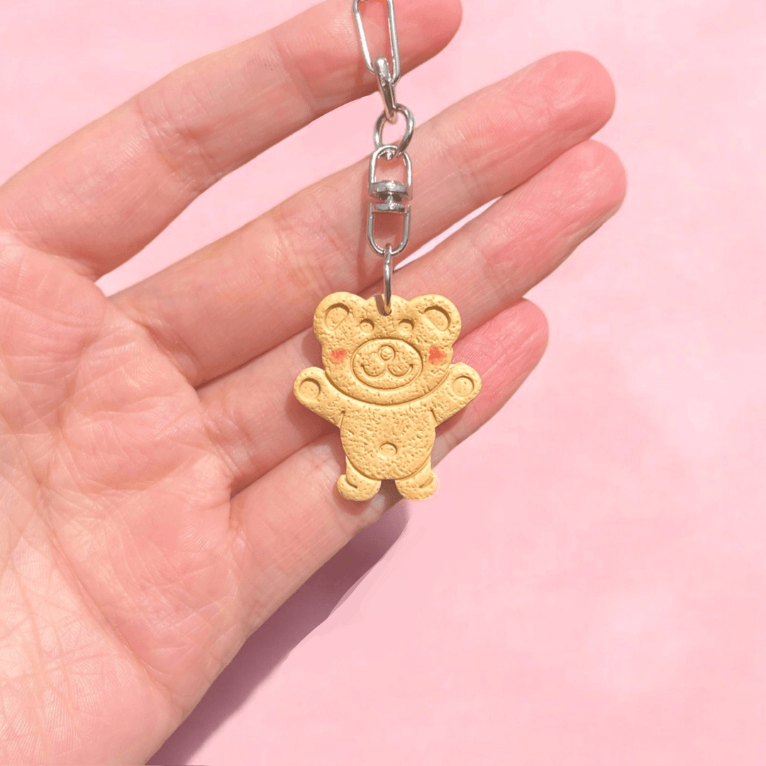 A hand holds the Tiny Teddy Polymer Clay Keyring, showcasing a small, beige teddy bear-shaped cookie with a smiling face and rosy cheeks. As part of New Zealand handmade gifts, it stands out against the soft pink background.