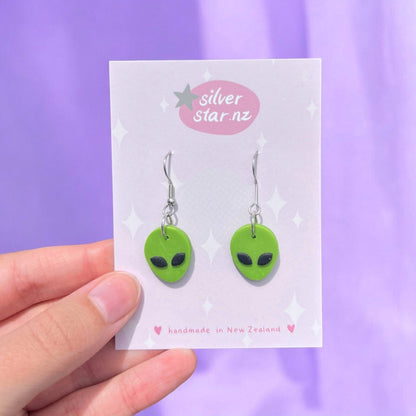 A hand displays a card featuring the Alien Polymer Clay Earrings, designed as handmade green alien heads with black eyes. The card carries the brand name silverstar.nz on a pink label at the top and mentions "handmade in New Zealand" below, set against a striking purple background.