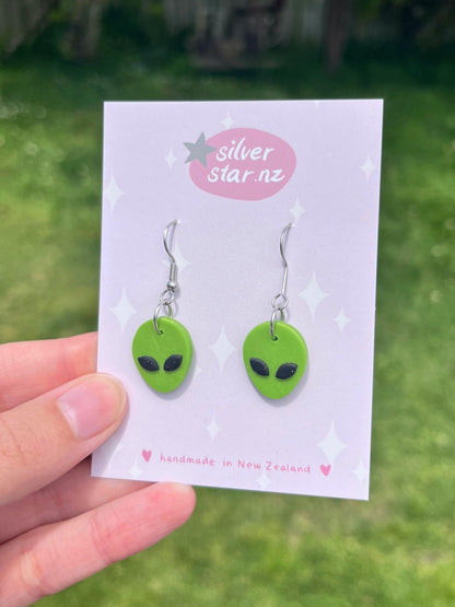 A hand holds a card showcasing the Alien Polymer Clay Earrings, featuring two green alien faces with striking black eyes. The card reads "silverstar.nz" and "handmade in New Zealand." Set against a grassy backdrop, these unique earrings beautifully capture both curiosity and craftsmanship.