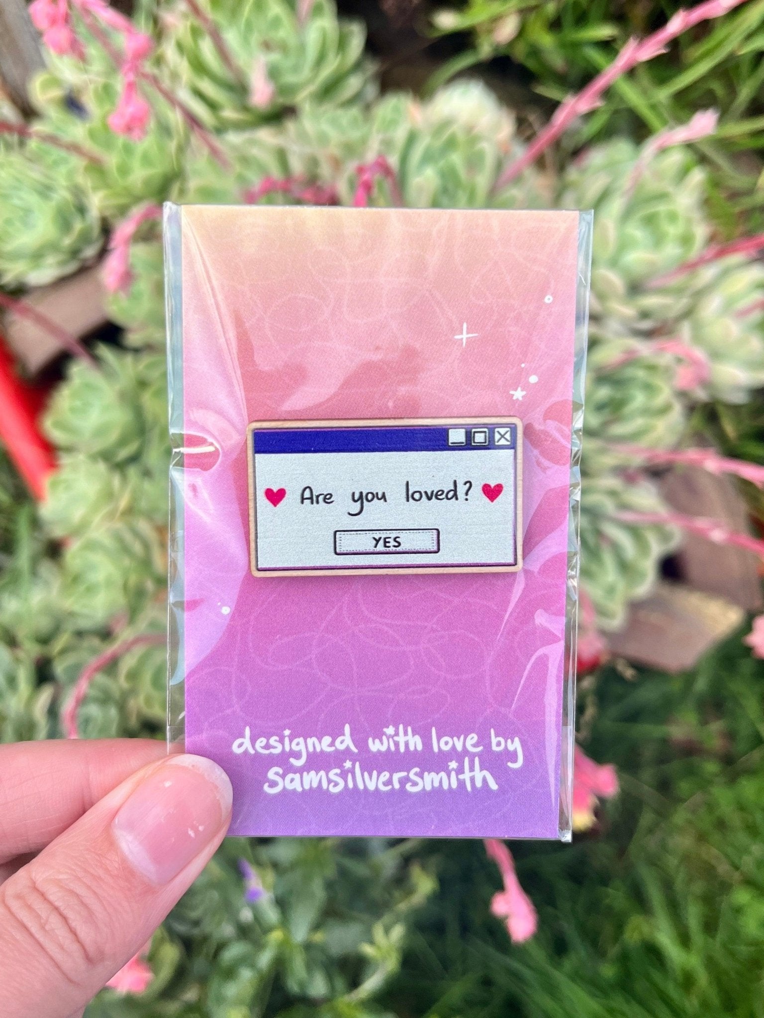 A hand holds a silverstar.nz wooden pin featuring an artist-designed digital pop-up window that asks, "Are you loved?" with a Yes option. "Designed with love by silverstar.nz" is printed on the card backing. Green plants and pink flowers blur in the background, enhancing its charm.