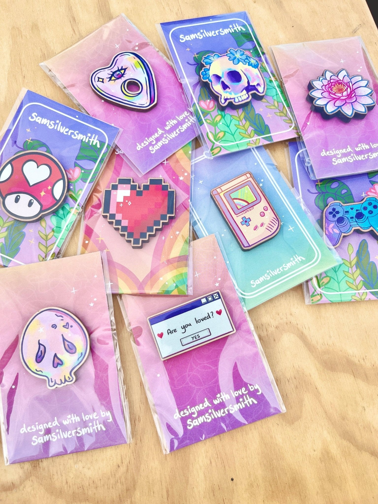 A collection of vibrant enamel pins, including artist-designed pieces such as a mushroom, heart, skull, flower, Gameboy, and gaming controller, is displayed on a cherry veneered MDF surface. Each pin is presented on a card featuring the inscription "designed with love by SamSilverSmith," with the standout piece being the Are You Loved Wooden Pin from silverstar.nz.