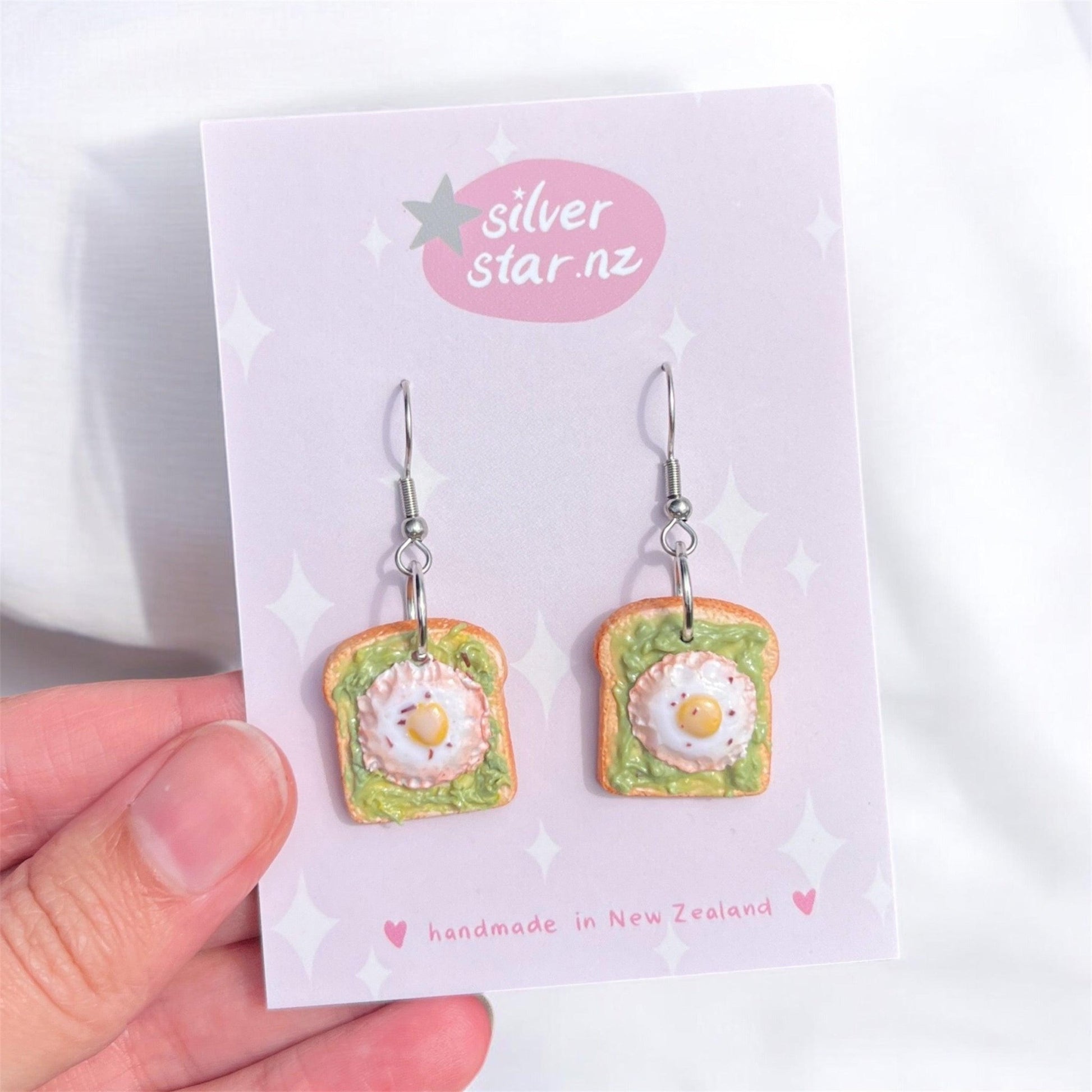 A hand displays a card featuring the Avocado and Egg on Toast Polymer Clay Earrings, styled as slices of bread with avocado and a sunny-side-up egg. The card, against a light backdrop, highlights "silverstar.nz" and accentuates "handmade in New Zealand.