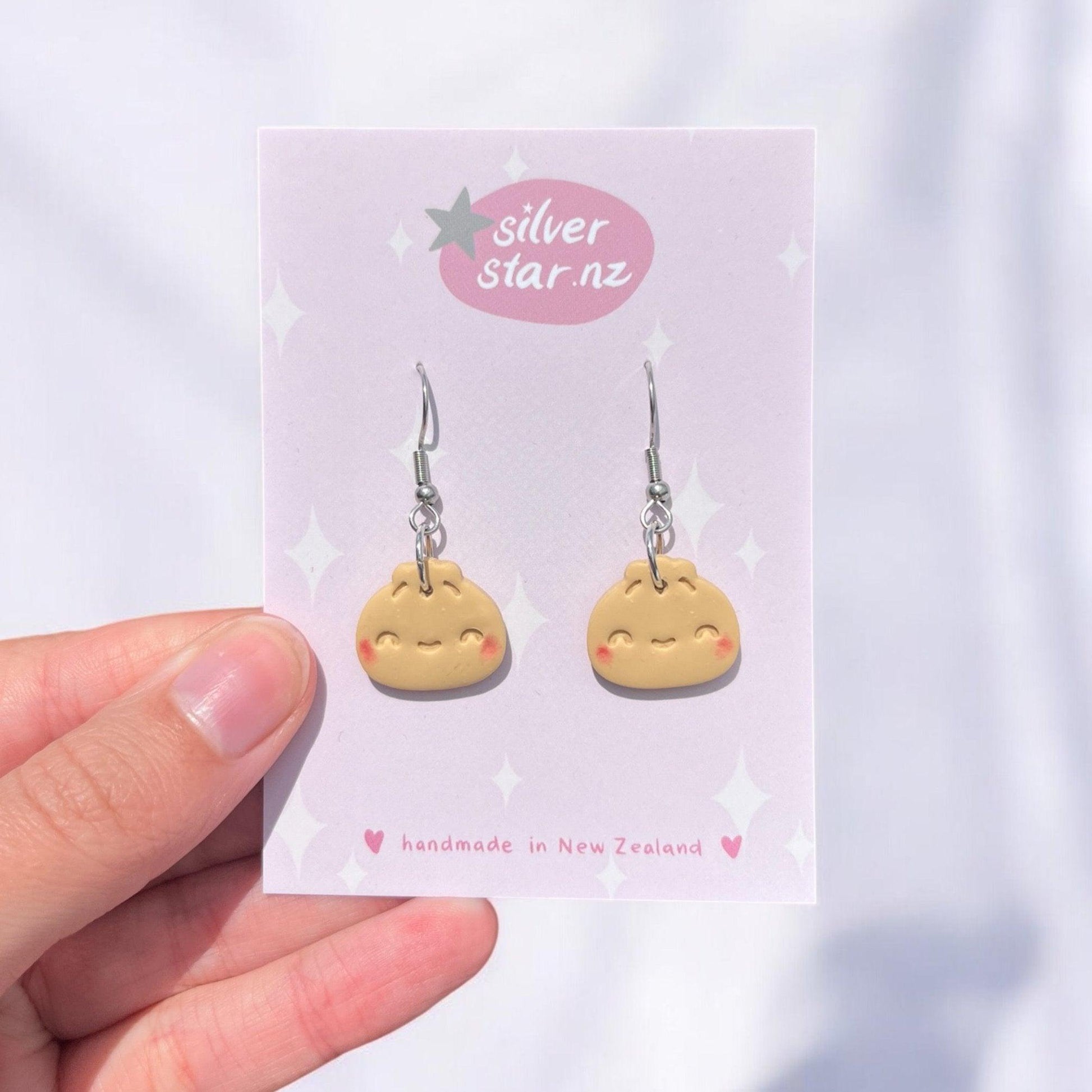 A hand holds a card to which Bao Bun Polymer Clay Earrings are attached. The earrings depict an adorable, smiling dumpling with rosy cheeks. The card displays "silverstar.nz" and "handmade in New Zealand," adorned with star decorations that emphasize the artisan craftsmanship. The background is a blurred white.