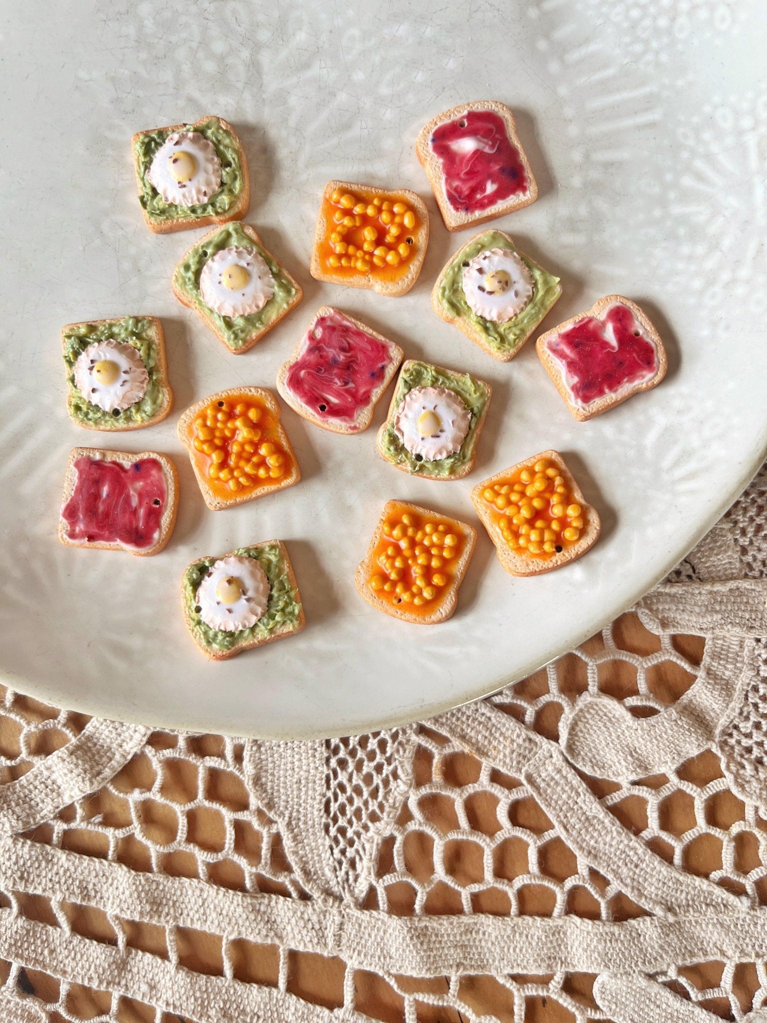 A white plate showcasing an assortment of vibrant, bite-sized snacks—pieces adorned with pink jam, green herb spread with white dollops, and yellow spheres—rests on a textured lace tablecloth. The whimsical design echoes the charm of the Beans on Toast Polymer Clay Earrings by silverstar.nz.