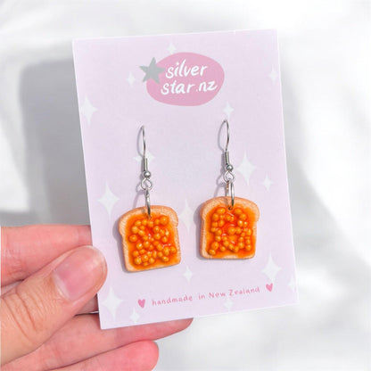 A hand displays the quirky Beans on Toast Polymer Clay Earrings, featuring miniature toast slices topped with orange beads that resemble baked beans. These handmade earrings are presented on a card decorated with stars and branded with silverstar.nz. Made in New Zealand, they come with stainless steel hooks for durability.