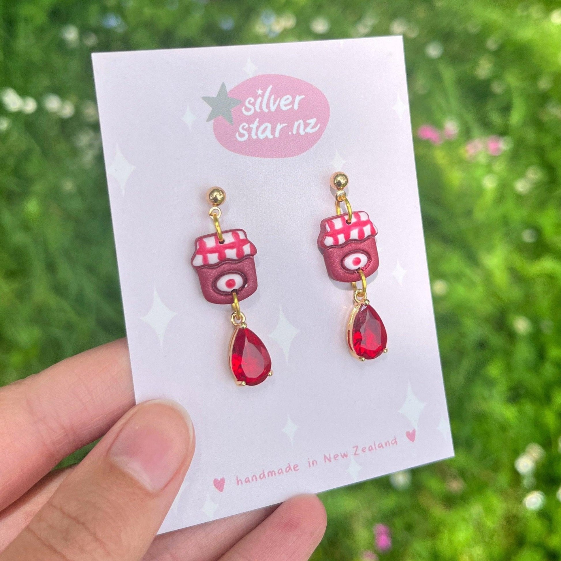 A close-up of a hand showcasing a pair of unique Berry Jam Jar Polymer Clay Earrings on a silverstar.nz card. These handmade beauties are adorned with red camera designs and teardrop-shaped red gems. The backdrop features blurred greenery, accentuating the charm of these earrings.