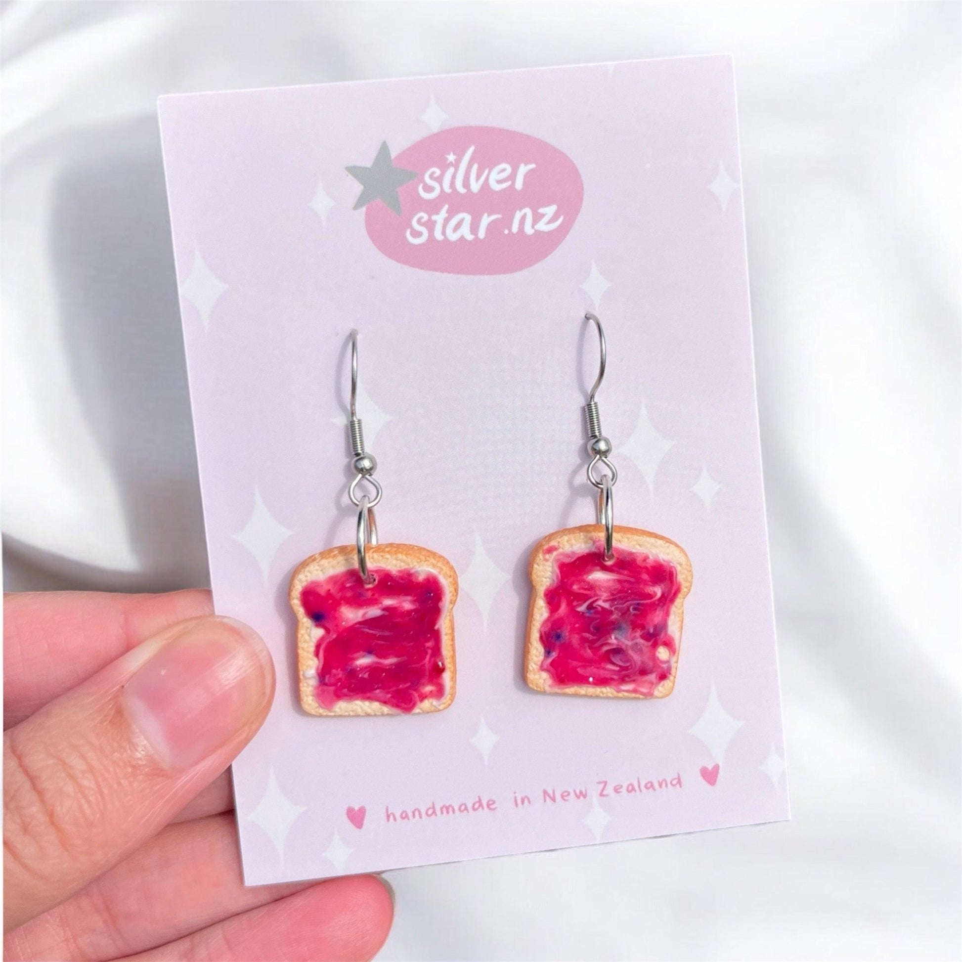 A hand holds a pair of whimsical Berry Jam on Toast Polymer Clay Earrings, elegantly displayed on a card. The card showcases the silverstar.nz logo and proudly states "handmade in New Zealand." These charming earrings feature stainless steel hooks.