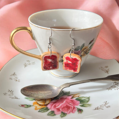 A porcelain teacup with floral designs holds the Berry Jam on Toast Polymer Clay Earrings by silverstar.nz, crafted to resemble slices of toast with red jam. It sits on a matching saucer adorned with a pink rose pattern, accompanied by a silver spoon. The earrings dangle elegantly from stainless steel hooks against the pink fabric backdrop.