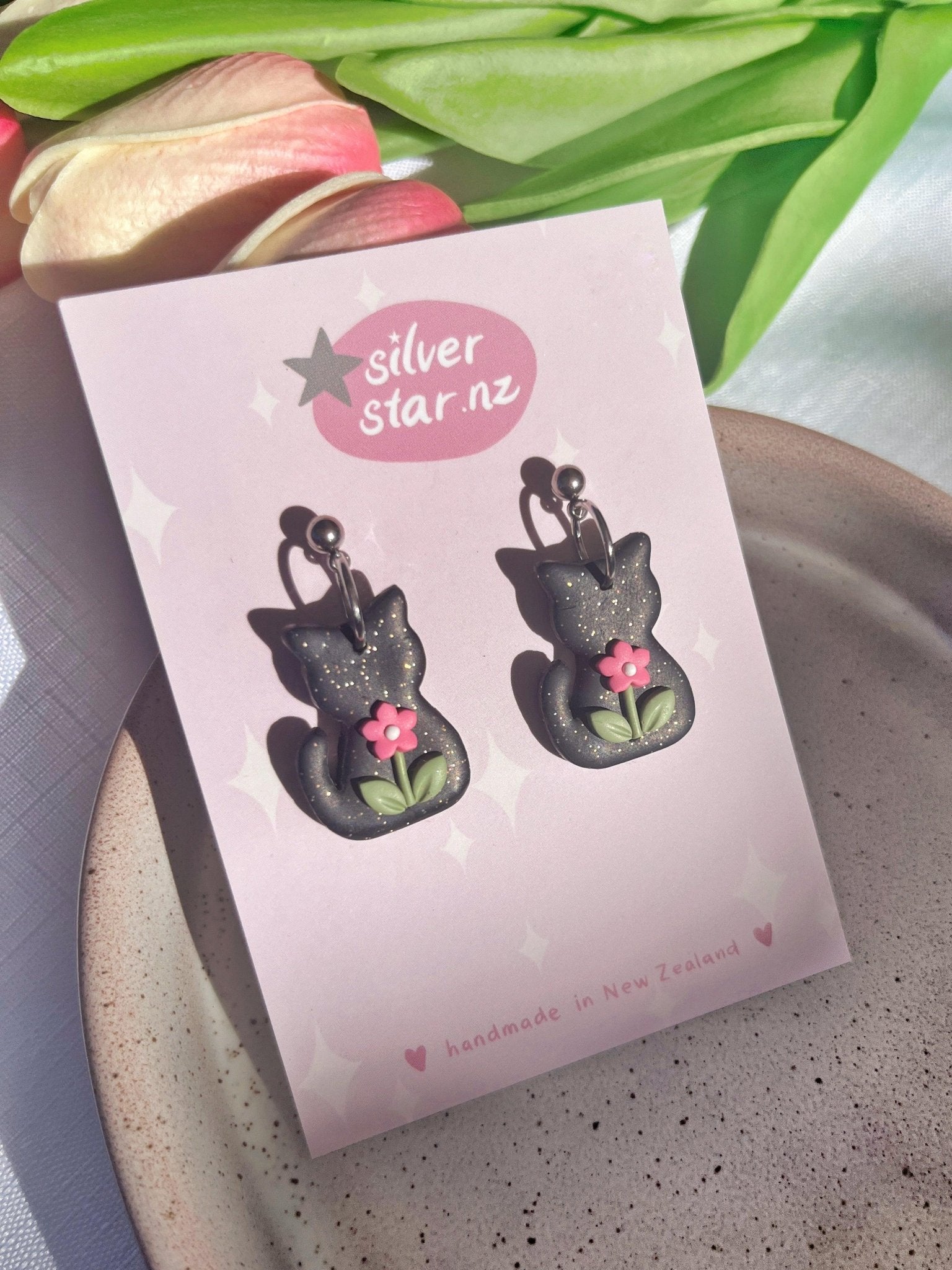 A close-up of silverstar.nz's Black Cat Polymer Clay Earrings showcases handmade earrings shaped like black cats with delicate pink flowers and green leaves. They are beautifully presented on a pink card. Sunlight casts gentle shadows, complemented by tulips and a textured ceramic plate, enhancing the charming scene.