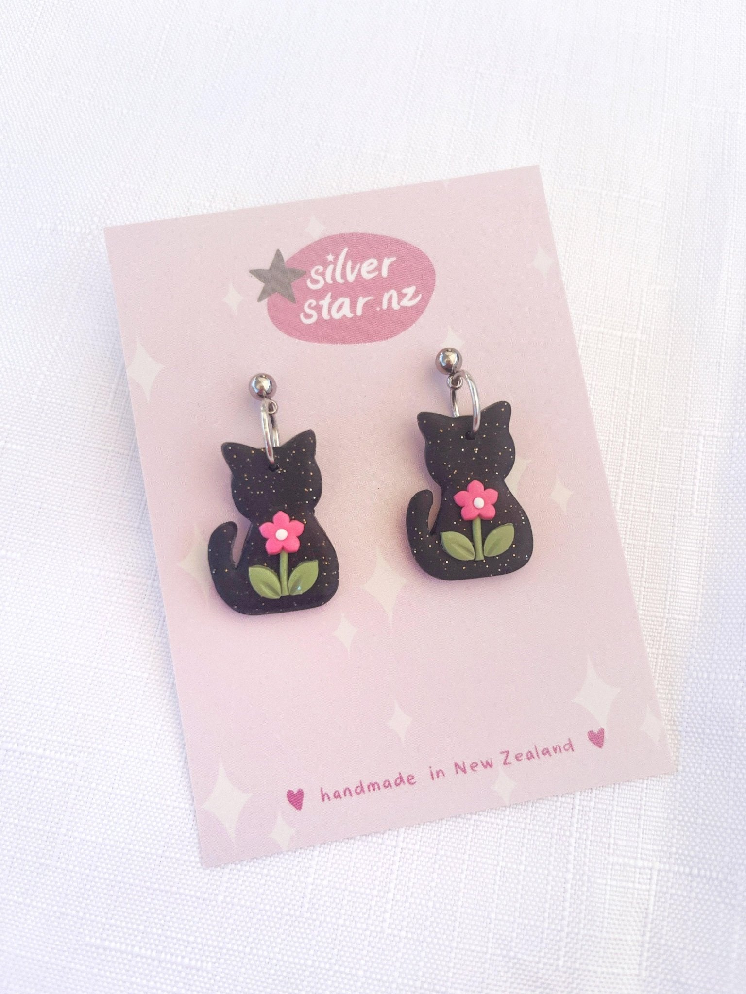 These delightful Black Cat Polymer Clay Earrings, featuring black cat designs embellished with pink flowers and green leaves, are meticulously handcrafted in New Zealand. They come with durable stainless steel hooks and are elegantly displayed on a card branded as silverstar.nz against a pristine white background.