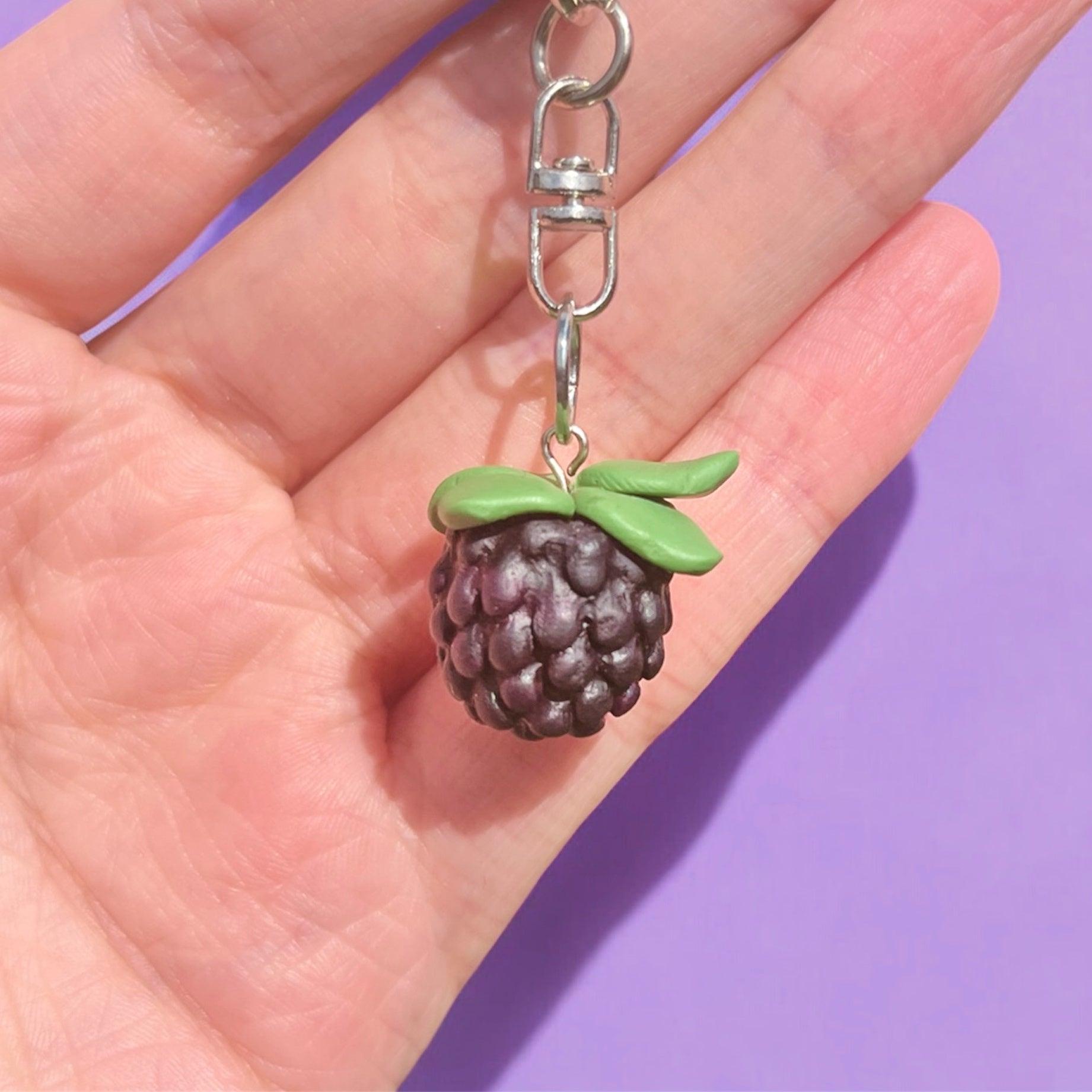 A hand holds the Blackberry Polymer Clay Keyring by silverstar.nz, featuring a uniquely designed blackberry shape crafted from durable polymer clay, with textured dark purple fruit and green leaves, set against a lavender background.