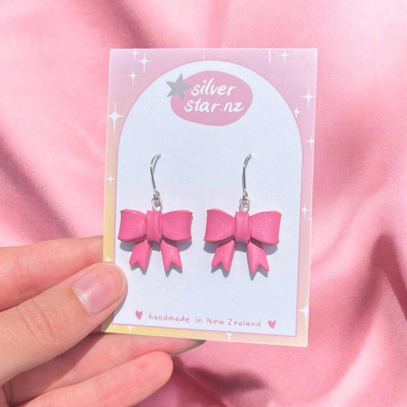 A hand presents the Bow Coquette Polymer Clay Earrings from silverstar.nz, delicately crafted into a pair of pink bows from handmade polymer clay. They are elegantly displayed on a card adorned with a pink and white starry design. The earrings, which feature stainless steel hooks, proudly bear the silverstar.nz label and are noted as handmade in New Zealand. A pink fabric serves as the backdrop.