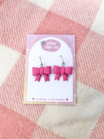 The Bow Coquette Polymer Clay Earrings from silverstar.nz showcase a charming design, with each pink bow-shaped earring meticulously handcrafted from polymer clay. They hang elegantly on stainless steel hooks, presented on a silverstar.nz card against a pink and cream checkered fabric, highlighting their distinct New Zealand craftsmanship.