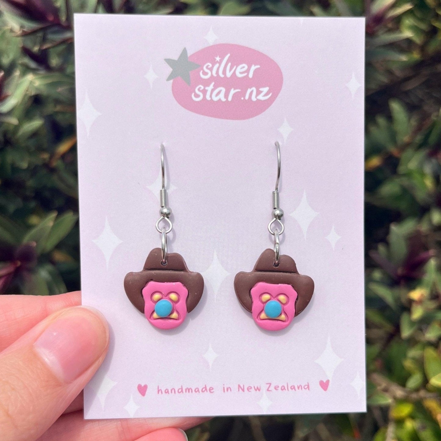 A close-up of a hand holding a card showcases handcrafted, colorful Bubble 'O Bill Polymer Clay Earrings. These intricate earrings feature pink faces and brown ears, dangling from stainless steel hooks. The card reads silverstar.nz and handmade in New Zealand, with blurred foliage in the background.