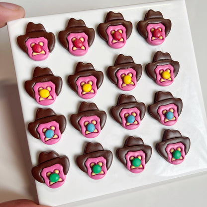 A grid of sixteen candies shaped like cowboy hats in chocolate, each adorned with a pink star badge at the center and showcasing a vibrant cowboy silhouette beneath, reminiscent of the Bubble 'O Bill Polymer Clay Earrings by silverstar.nz. The candies are neatly arranged on a white square surface.