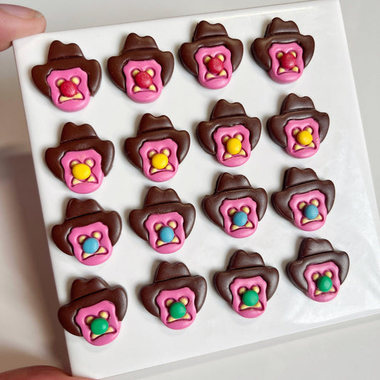 A grid of sixteen candies shaped like cowboy hats in chocolate, each adorned with a pink star badge at the center and showcasing a vibrant cowboy silhouette beneath, reminiscent of the Bubble 'O Bill Polymer Clay Earrings by silverstar.nz. The candies are neatly arranged on a white square surface.