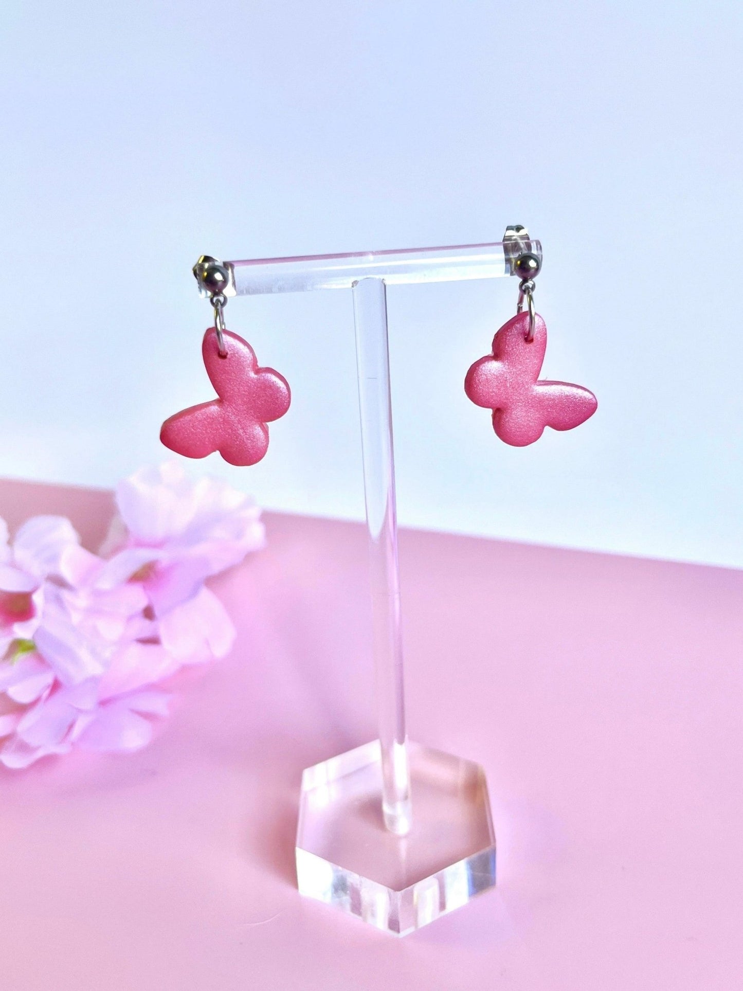 The Butterfly Polymer Clay Earrings from silverstar.nz, with their distinctive pink butterfly shape, are showcased elegantly on a clear jewelry stand. Soft pink flowers adorn the background while the handmade earrings rest beautifully on a pink surface.