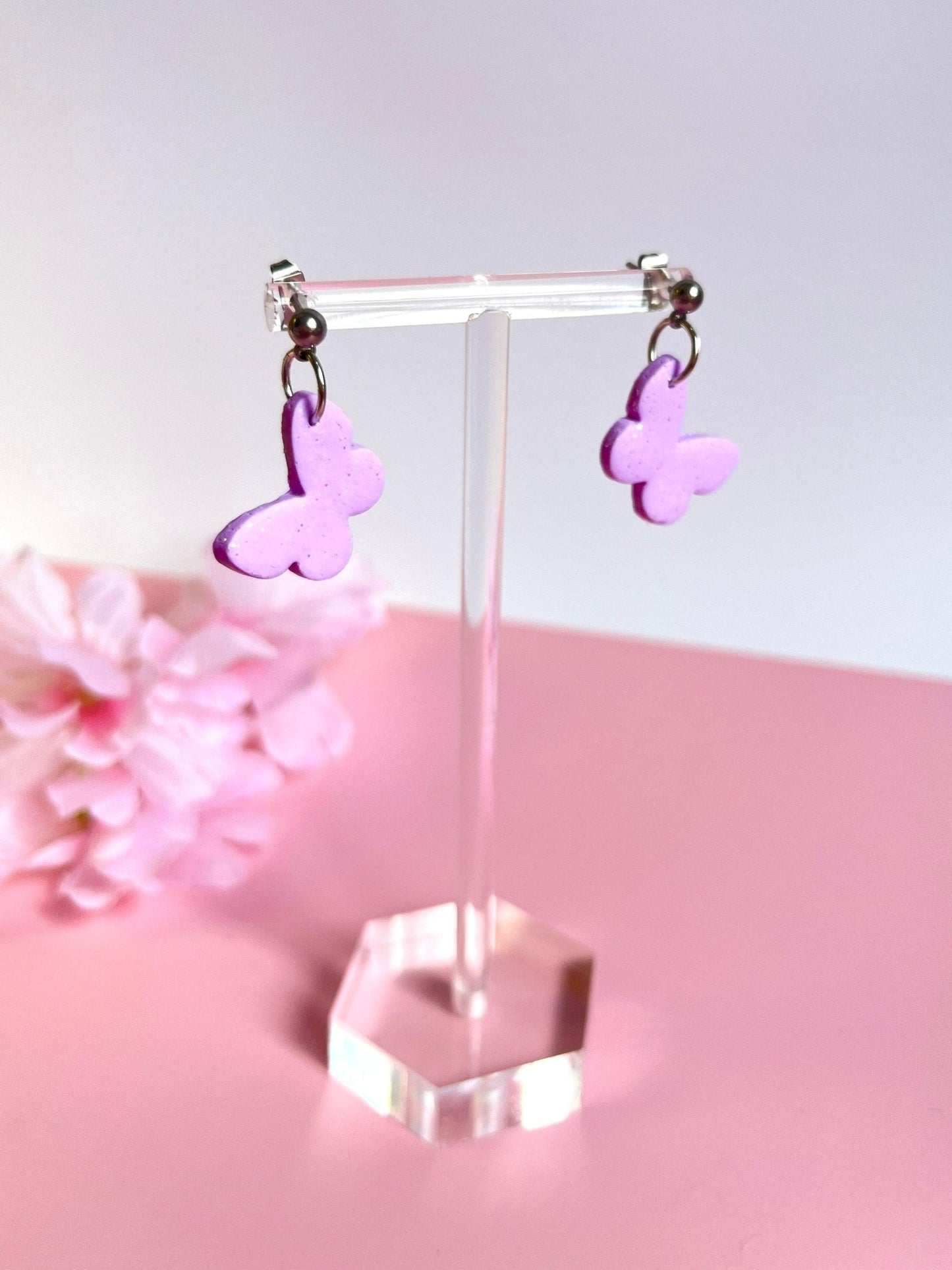 The Butterfly Polymer Clay Earrings by silverstar.nz are showcased on a clear stand against a soft pink backdrop, with delicate pink flowers softly blurred in the corner. These unique handmade earrings add an artistic flair to any outfit.