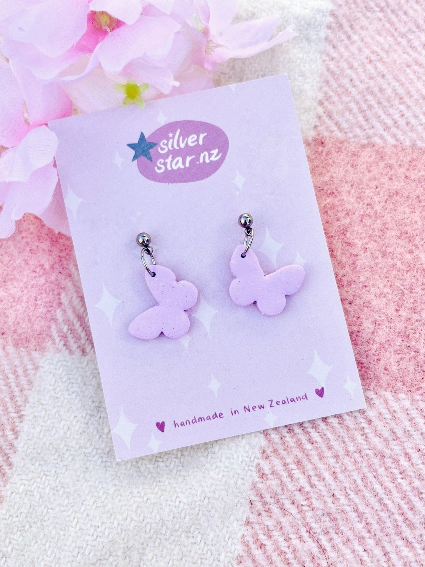 The Butterfly Polymer Clay Earrings come on a card featuring the silverstar.nz logo. The backdrop is a gentle pink and white checkered design, accented with delicate light pink flowers. These one-of-a-kind earrings are handcrafted in New Zealand from premium polymer clay.