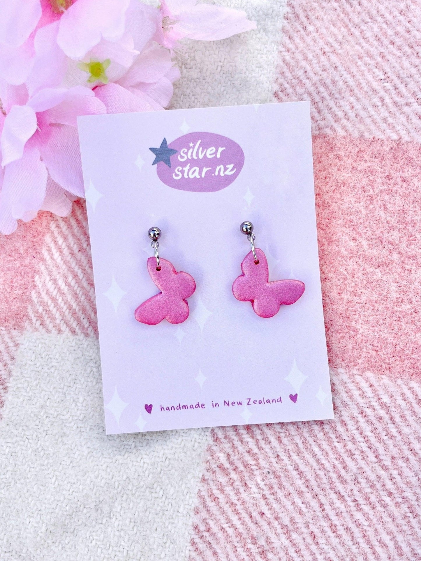 Butterfly Polymer Clay Earrings by silverstar.nz feature pink butterfly shapes crafted from polymer clay, displayed on a floral background card. The card's starry design includes the message "handmade in New Zealand," adding to the charm and uniqueness of these delightful accessory pieces.