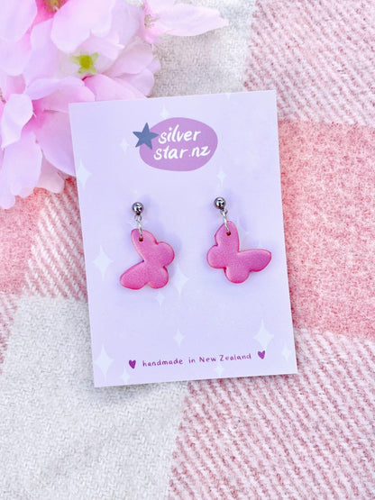 Butterfly Polymer Clay Earrings by silverstar.nz feature pink butterfly shapes crafted from polymer clay, displayed on a floral background card. The card's starry design includes the message "handmade in New Zealand," adding to the charm and uniqueness of these delightful accessory pieces.