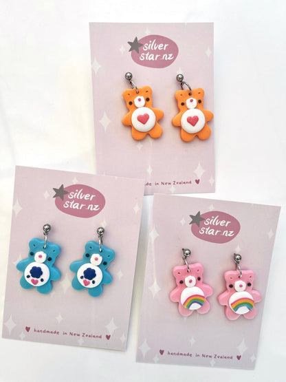 A collection of three pairs of Care Bear-themed polymer clay earrings, presented on display cards, highlights their unique designs. This vibrant assortment includes a pair in orange with heart motifs, a blue pair with paw prints, and a pink pair adorned with rainbows. The cards are labeled "handmade in New Zealand by silverstar.nz.